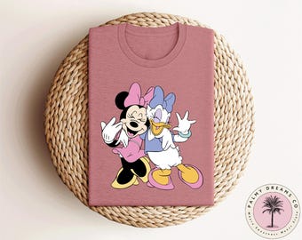 May include: A pink t-shirt with a graphic of Mickey Mouse and Daisy Duck. Mickey is wearing a red and white dress and Daisy is wearing a blue and white dress.