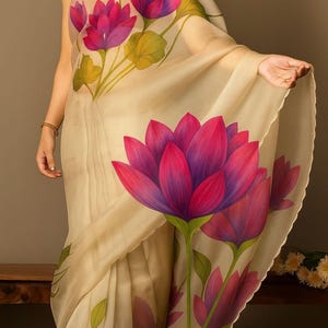 May include: A cream-coloured saree featuring large, vibrant pink and purple lotus flower designs with green leaves. The sheer fabric drapes elegantly, showcasing the floral pattern. The saree is a traditional garment.