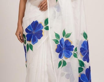 May include: White saree with a halter top, adorned with large, hand-painted blue flowers with gold accents and green leaves. The floral pattern is repeated along the border of the sheer fabric, creating a flowing, elegant look.
