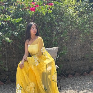 Op de afbeelding: Een vrouw draagt een doorschijnende gele sari met witte bloemmotieven en groene bladeren. De sari is over een witte top gedrapeerd. De achtergrond omvat groen gebladerte en roze bloemen.