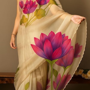 May include: A cream-coloured sari featuring large, vibrant pink and purple lotus flower designs with green leaves. The floral pattern is hand-painted on the sheer fabric. The sari is draped, showcasing the intricate artwork and flowing material.