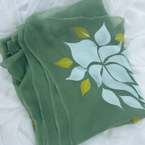 May include: A green sheer scarf with a white and yellow floral design. The scarf is folded and lying on a white surface.