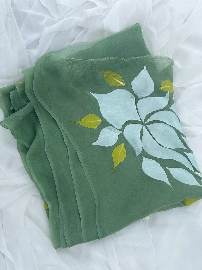 May include: A green sheer scarf with a white and yellow floral design. The scarf is folded and lying on a white surface.