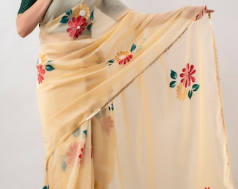 May include: A sheer, pale yellow saree with a floral pattern in red, yellow, and green. The saree has a gold sequined border. The model is wearing a dark green top underneath the saree.