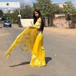 Op de afbeelding: Een heldergele outfit met witte bloemmotieven. De set bestaat uit een lange rok en een doorschijnende sjaal. Het bloemmotief wordt herhaald op de mouwen en het lijfje. De outfit wordt buiten gedragen op een zonnige dag.