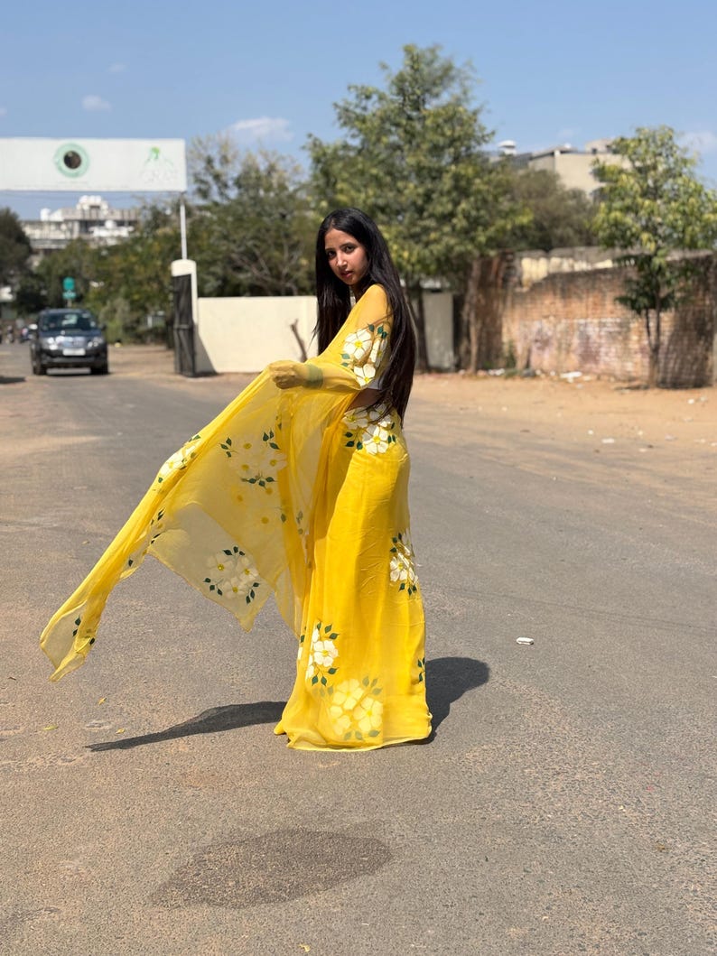 Op de afbeelding: Een heldergele outfit met witte bloemmotieven. De set bestaat uit een lange rok en een doorschijnende sjaal. Het bloemmotief wordt herhaald op de mouwen en het lijfje. De outfit wordt buiten gedragen op een zonnige dag.