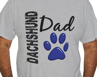 May include: A light gray t-shirt with the words "DACHSHUND Dad" printed on the front. The word "DACHSHUND" is in a vertical, distressed font, and "Dad" is in a cursive font. A blue paw print is below the word "Dad".