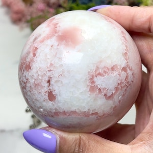 May include: A sphere-shaped crystal with a mix of white and pink hues. The crystal ball has a textured surface with a cracked appearance, and the pink color is distributed throughout the white base. The sphere is held in a hand.