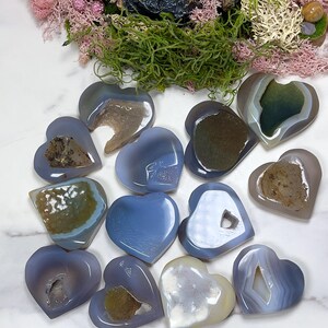 May include: A collection of heart-shaped agate stones in shades of blue, gray, and white. Some stones have visible crystal formations and unique patterns. The arrangement is on a white surface, with green moss and pink foliage in the background.