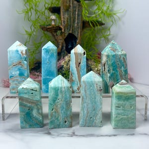 May include: Eight polished gemstone towers in shades of blue and green, with brown veining. The crystals are displayed on a clear acrylic shelf, with a decorative background of greenery and a small wooden structure.