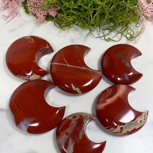 May include: Six polished red jasper crescent moon shapes with white and tan veining. The gemstones are arranged on a white surface with green and pink decorative moss in the background. Each moon measures approximately 2 inches across.