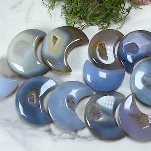 May include: A collection of polished agate crescent moon shapes in various shades of blue, gray, and brown. Some moons have a geode center. The arrangement includes full and half-moon designs, showcasing natural stone formations.