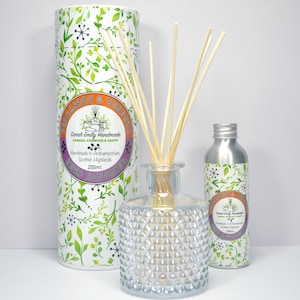 May include: A clear glass bottle with a textured surface filled with a reed diffuser. The bottle is surrounded by a silver bottle and a cardboard tube. The tube has a white label with the text "Sweet Emily Handmade Natural Cosmetics & Crafts Handmade in Ardnamurchan Scottish Highlands Reed Diffuser 200ml" and a floral design. The silver bottle has a similar label with the same floral design.