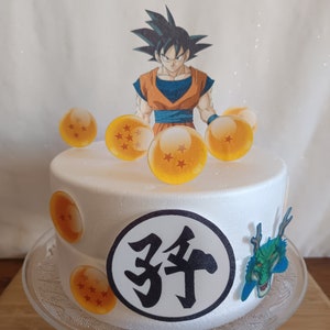 May include: A white cake decorated with a cartoon image of Goku from Dragon Ball Z, surrounded by orange dragon balls and a black circle with the Japanese character for "year" in it. The cake is on a clear glass cake stand.