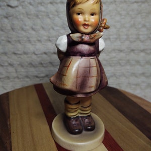 May include: A porcelain figurine of a girl wearing a plaid dress and a white and red bonnet. She has brown hair and brown eyes. The figurine is standing on a round base.