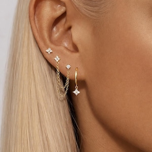 May include: A close-up of a person's ear adorned with multiple gold earrings. The earrings include small stud earrings, a double chain earring, and a hoop earring. Each earring features a floral design with small, sparkling stones.