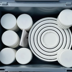 Puede incluir: Un contenedor de almacenamiento gris lleno de objetos circulares blancos. Se ven varias pilas de objetos cilíndricos, junto con un conjunto de anillos concéntricos con rayas negras. También hay un pequeño folleto, lo que sugiere un conjunto de productos.