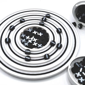 May include: A white circular tray with concentric black rings, representing an atom's electron shells. Black and white round beads with plus and minus signs are arranged in the center and on the rings, simulating protons, neutrons, and electrons.