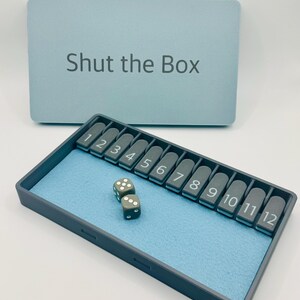 May include: A blue Shut the Box game with a gray tray and two gray dice. The tray has numbers 1 through 12.