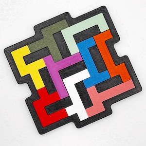 Op de afbeelding: Een zwart puzzelbord met verschillende gekleurde puzzelstukken. De stukken zijn L-vormig en T-vormig en passen in elkaar om een groter ontwerp te creëren.