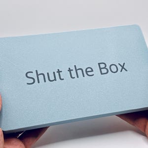 May include: A light blue rectangular box with the text "Shut the Box" printed on the top.