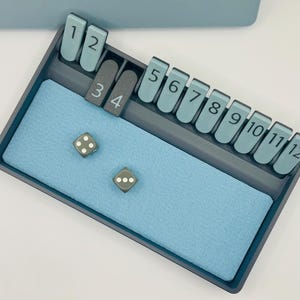 May include: A gray game tray with a blue felt lining. The tray has slots for numbered tiles, from 1 to 12. Two gray dice with white dots are on the felt lining.