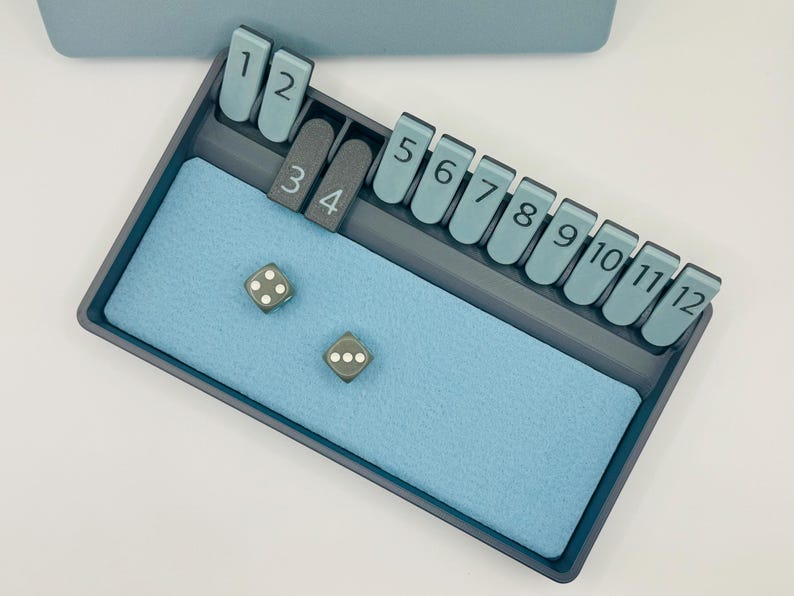 May include: A gray game tray with a blue felt lining. The tray has slots for numbered tiles, from 1 to 12. Two gray dice with white dots are on the felt lining.