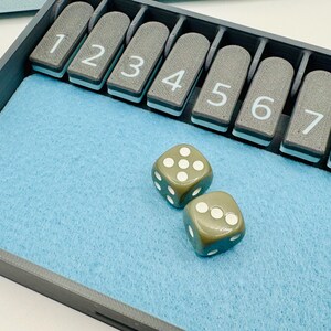 May include: A gray tray with a blue felt lining and a row of numbered tiles from 1 to 8. Two green dice with white dots are on the felt.