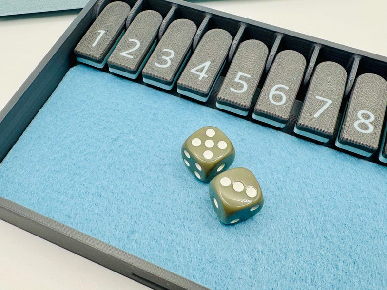 May include: A gray tray with a blue felt lining and a row of numbered tiles from 1 to 8. Two green dice with white dots are on the felt.