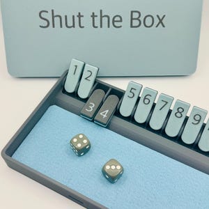 May include: A blue Shut the Box game with the lid open, showing the numbered tiles and two dice. The dice are grey with white dots.
