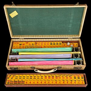 May include: A vintage Mah Jongg set in a textured beige carrying case with a green interior. The case contains yellow tiles with red and black symbols, four colorful racks in green, yellow, purple, and blue, and a separate tray of yellow tiles.