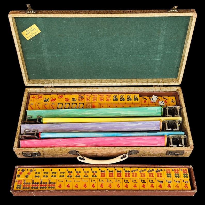 May include: A vintage Mah Jongg set in a textured beige carrying case with a green interior. The case contains yellow tiles with red and black symbols, four colorful racks in green, yellow, purple, and blue, and a separate tray of yellow tiles.