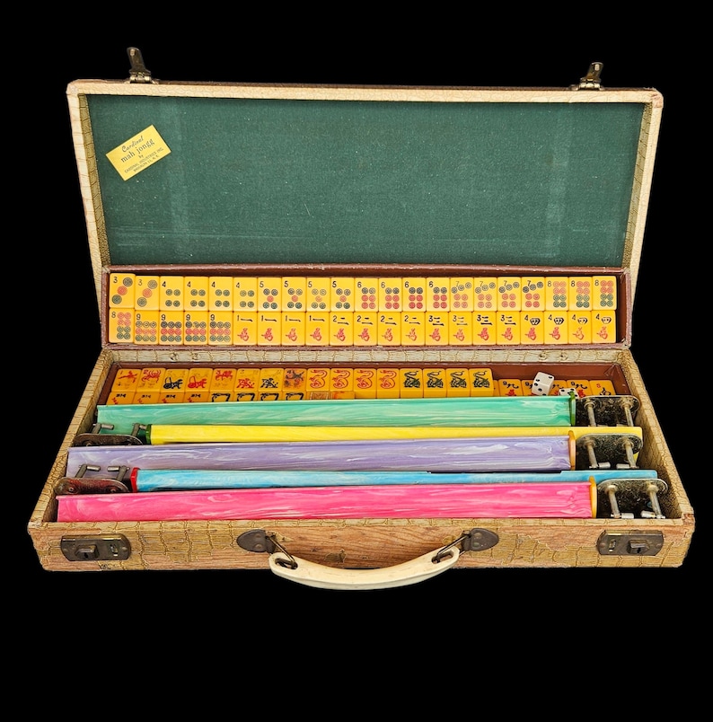 May include: A vintage Mah Jongg game set housed in a textured tan carrying case with a green interior lining. The set includes yellow tiles with red, black, and green markings, along with four colorful racks in pink, blue, purple, and yellow.