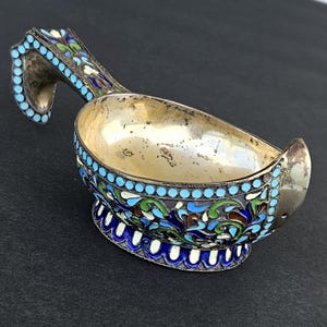 Op de afbeelding: Een zilverkleurige, sierlijke lepel met een gebogen handvat en een komvormige schep. De lepel is versierd met blauwe, groene, witte en bruine emaille bloemmotieven en turquoise stippen. De lepel heeft een decoratieve basis.
