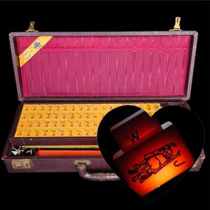 Può includere: Set di Mahjong vintage in una valigetta marrone con interno rosso. Il set include tessere gialle con segni rossi, un bastoncino di legno e un piccolo cassetto. La valigetta è aperta, rivelando le tessere e gli accessori.