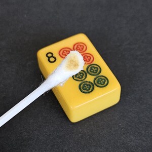 May include: A yellow mahjong tile featuring the number 8 and a pattern of six circular symbols in red and green. A white cotton swab with a brown residue on its tip is positioned diagonally across the surface of the tile for cleaning purposes.