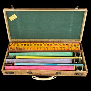 May include: A vintage Mah Jongg game set in a textured tan carrying case with a green interior lining. The set includes yellow tiles with red and black markings, and four colorful tile racks in green, yellow, purple, blue, and pink.