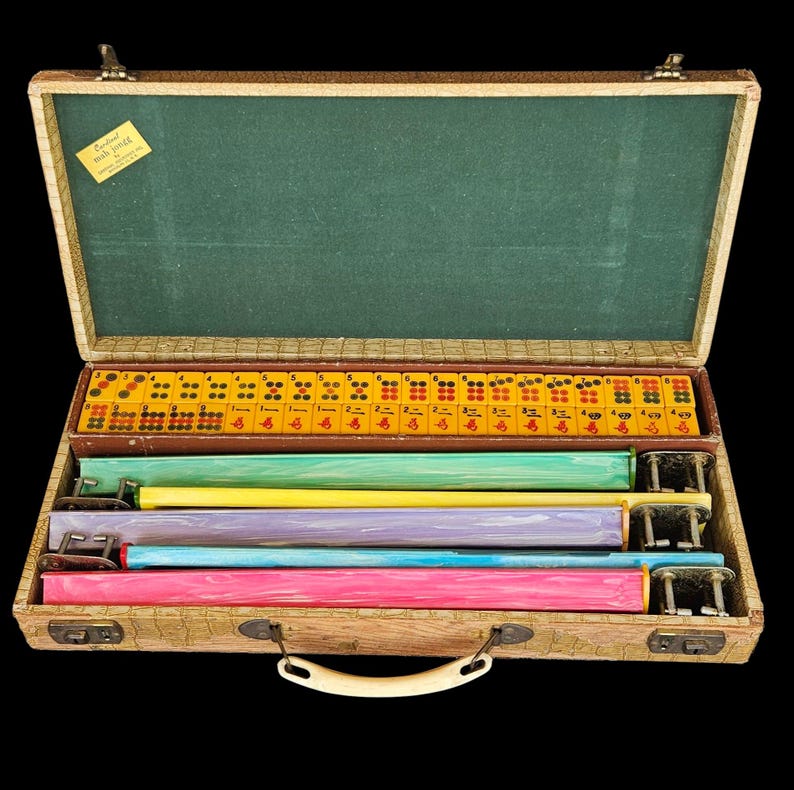 May include: A vintage Mah Jongg game set in a textured tan carrying case with a green interior lining. The set includes yellow tiles with red and black markings, and four colorful tile racks in green, yellow, purple, blue, and pink.