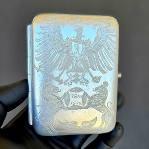 May include: Silver cigarette case with engraved details. The case features an eagle crest, lions, and the year 1914. The case is rectangular with a hinged lid and a small clasp on the side.