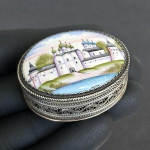 May include: Oval silver trinket box with a hand-painted miniature landscape scene. The lid features a detailed depiction of a white building with green and blue domes, set against a pastel sky. The box has a filigree design.