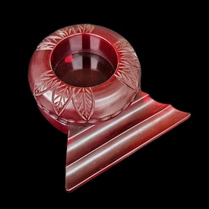 Puede incluir: Un cenicero circular de color rojo intenso con un patrón de hojas talladas alrededor del cuenco central. El objeto incluye una zona de descanso amplia, plana y extendida con dos ranuras paralelas, hecho de un material sólido, liso y de color carmesí oscuro.