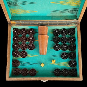 Puede incluir: Tablero de backgammon abierto con fichas marrones y negras, dos cubiletes de dados de cuero y dos dados amarillos. El tablero tiene una superficie de juego turquesa con detalles dorados y negros, y un marco de madera.