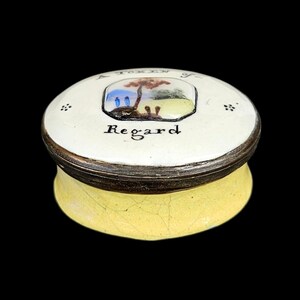 May include: A round ceramic trinket box with a cream-colored lid featuring a hand-painted landscape scene, the text A TOKEN OF Regard, and small floral accents. The base of the box is yellow with a visible crackle glaze finish and a dark rim.