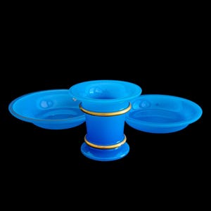 May include: A set of three blue glass pieces: a vase and two matching bowls. The vase has a cylindrical shape with gold bands. The bowls are shallow with a gold rim. All pieces are against a black background.