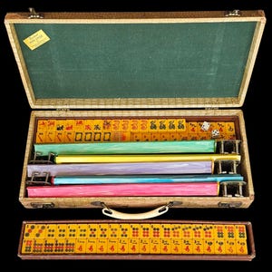 May include: A vintage Mah Jongg game set in a textured beige carrying case with a green interior lining. The case contains yellow tiles with red and black symbols, four colorful tile racks in green, yellow, purple, blue, and pink, and two dice.