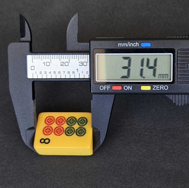 May include: A yellow mahjong tile featuring four red circular symbols and four green circular symbols, with the number 8 printed in black. A black digital caliper is measuring the tile, showing a width of 31.4 mm on its clear digital display screen.