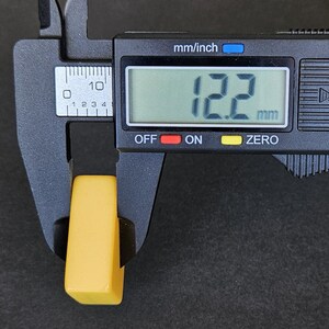 May include: A small, rectangular yellow block being measured by a black digital caliper. The digital display clearly shows a measurement of 12.2 mm. The object has a smooth, solid yellow surface and is positioned between the jaws of the measuring tool.