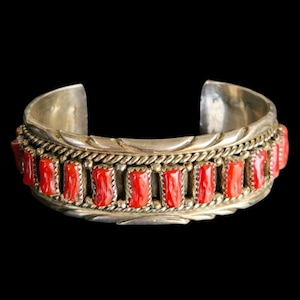 May include: A silver cuff bracelet featuring a line of rectangular, vibrant red stones. The bracelet has an intricate design with a braided rope pattern and decorative edges. The background is a solid black.