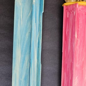 May include: Two vertical rectangular plastic dispensers with a marbled finish. One dispenser is light blue with white swirls, and the other is bright pink with white swirls. Both feature a yellow cap at the top and a grooved, channeled design.
