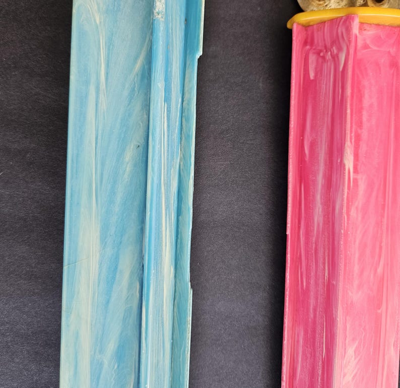 May include: Two vertical rectangular plastic dispensers with a marbled finish. One dispenser is light blue with white swirls, and the other is bright pink with white swirls. Both feature a yellow cap at the top and a grooved, channeled design.